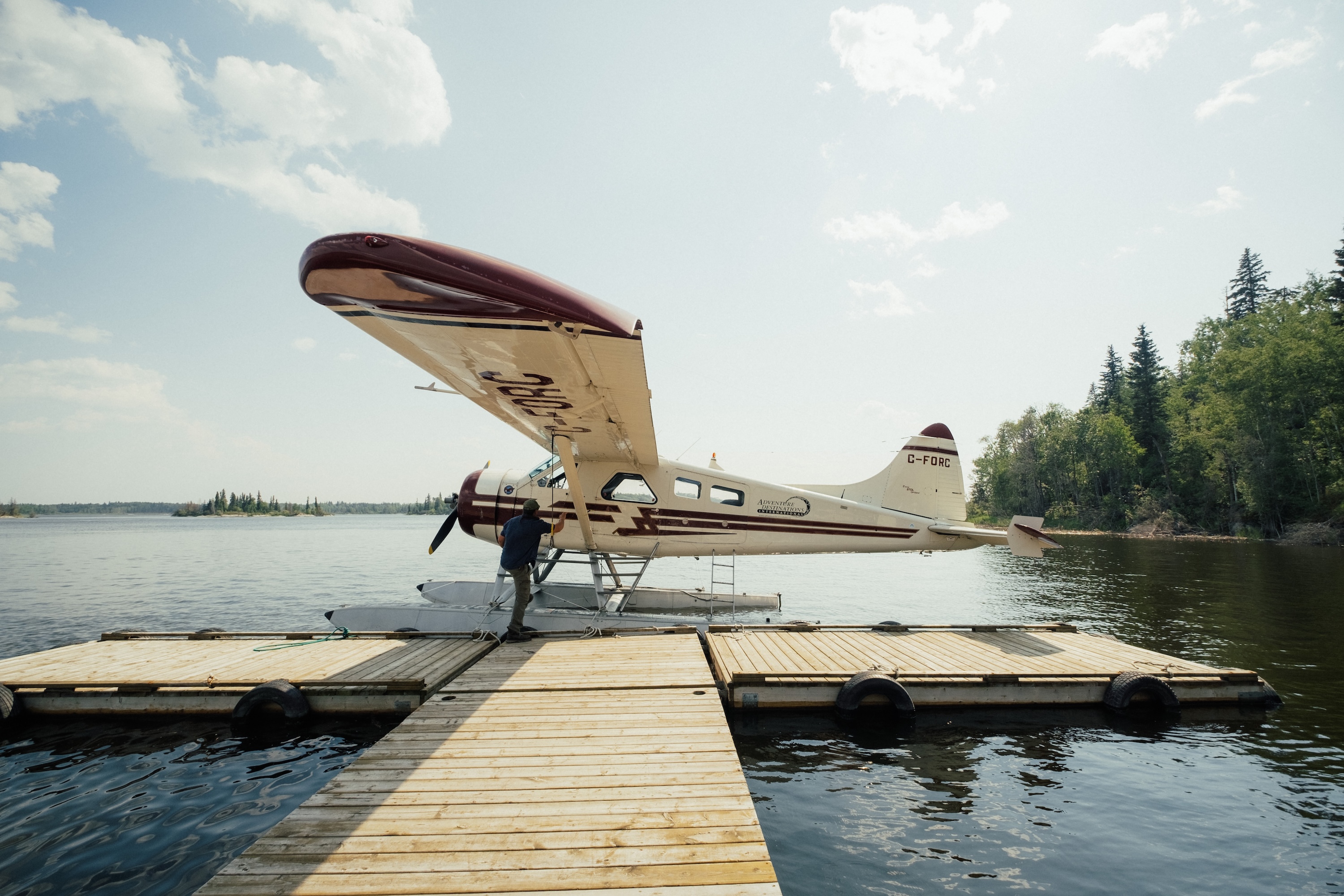 A white and maroon floatplane docked at a wooden pier on a calm lake surrounded by evergreen trees.