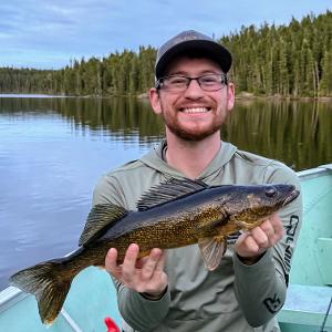 A smiling man holding a large walleye while sitting in a boat on a calm northern lake with a pine forest reflected in the background.