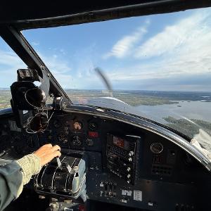 Interior of a bush plane cockpit showing the instrument panel and a view of lakes and forests through the windshield.