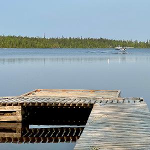 A wooden dock in the foreground of a calm lake with a floatplane taxiing in the distance against a backdrop of evergreen trees.
