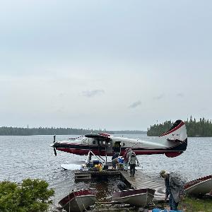 A red and white Otter floatplane docked at a wooden pier on Elbow Lake with several people loading gear.