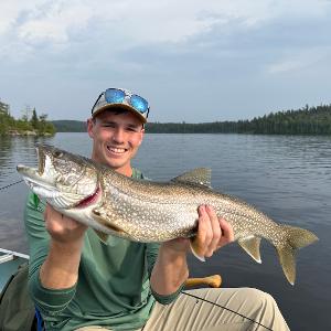 A smiling young man in a canoe holding a large Lake Trout horizontally against a backdrop of a calm lake and pine forest.