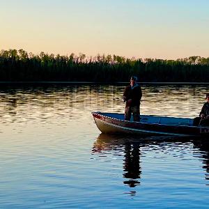 Two fishermen in a Lund boat on a calm lake at sunset with a forested shoreline in the background.