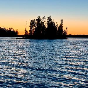 A wide shot of a wind-ruffled lake at sunset with a small island of pine trees silhouetted against an orange sky.