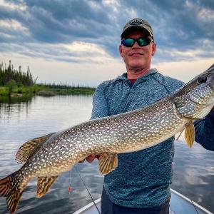 A fisherman in a boat holding a large, spotted Northern Pike horizontally against a backdrop of a blue lake and sky.